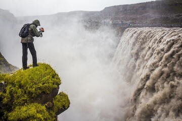 A man with backpack and camera is taking photo of impressive Dettifoss waterfall in Iceland. The falls are 100 metres wide and have a drop of 44 metres down to the canyon.