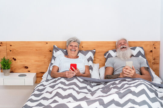 Happy senior couple watching videos with mobile phones while laughing together - Mature people having fun using smartphones at home - Technology, joyful elderly lifestyle - Focus on faces - Powered by Adobe