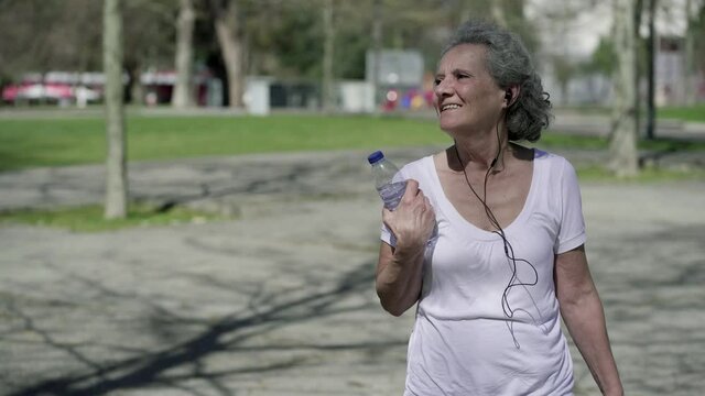 Happy elderly woman in earphones walking in park, holding bottle of water, listening to music, Medium shot, front view. Outdoor activity or leisure concept