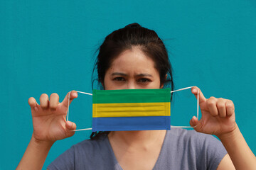 A woman with Gabon flag on hygienic mask in her hand and lifted up the front face on light blue color background. Tiny Particle or virus corona or Covid 19 protection.