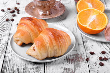 Two croissants of yeast dough on ceramic plate, freshly brewed coffee in a cup and oranges on white wooden table. Breakfast table setting