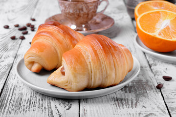 Two croissants of yeast dough on ceramic plate, freshly brewed coffee in a cup and oranges on white wooden table. Breakfast table setting