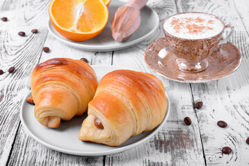 Two croissants of yeast dough on ceramic plate, freshly brewed coffee in a cup and oranges on white wooden table. Breakfast table setting