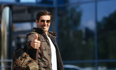 Portrait of happy satisfied handsome male tourist in sunglasses and casual jacket, carrying backpack looking at camera and showing thmb up
