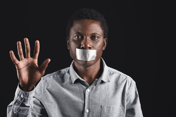 Sad African-American man with taped mouth showing stop gesture on dark background. Stop racism