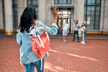 A cute african american girl student with a pink backpack waving to a group of students near the...