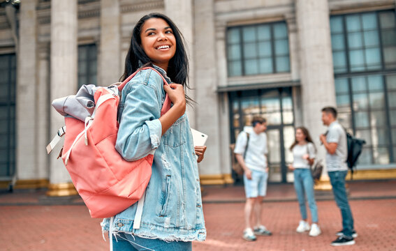 A cute African American girl student with a pink backpack and a laptop on the background of a group of students near the campus.