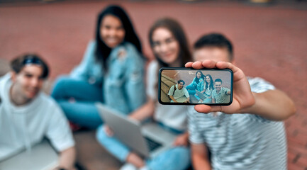 A group of students are sitting on the steps near the campus with laptops, relaxing, chatting and taking selfies on a smartphone.