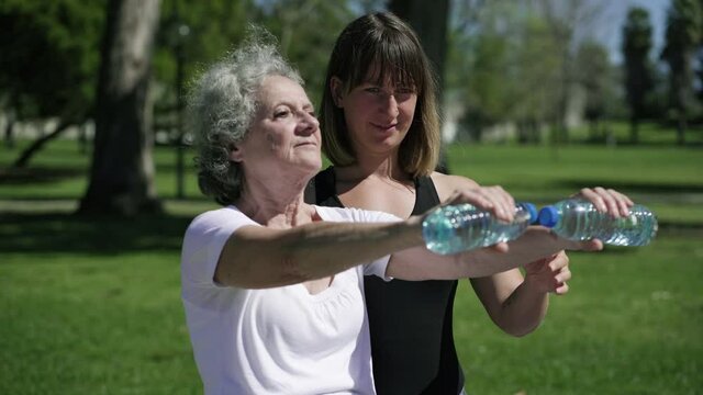 Focused old lady lifting weight and training hand muscles with instructor in park. Medium shot. Fitness for senior or sport activity concept