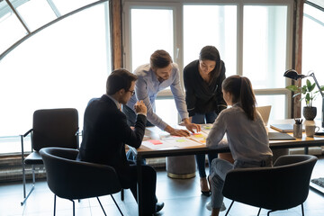 Concentrated diverse businesspeople gather at desk in office work together analyzing company paperwork, colleagues coworkers brainstorm discuss financial documents at meeting, cooperation concept
