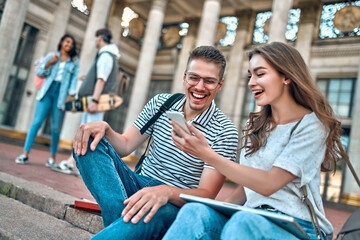 A couple of students with backpacks and a laptop sit on the steps near the campus. The girl shows the guy something on her smartphone.