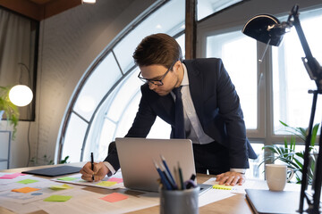 Naklejka premium Concentrated caucasian young businessman in formal suit handwrite prepare company paper statistics or report, focused male employee busy working on laptop, analyzing documents or paperwork in office