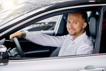 smiling handsome man driving a car, he sits inside of his new auto, male made purchase in car showroom