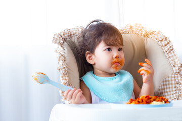 Adorable hungry toddler girl or infant baby eating delicious spaghetti food with tomato sauce on...
