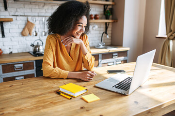 Fototapeta A cheerful mixed-race girl uses laptop for remote work or home leisure while sitting in the kitchen at home. Side view a nice girl with an afro hairstyle looks at screen with a smile obraz