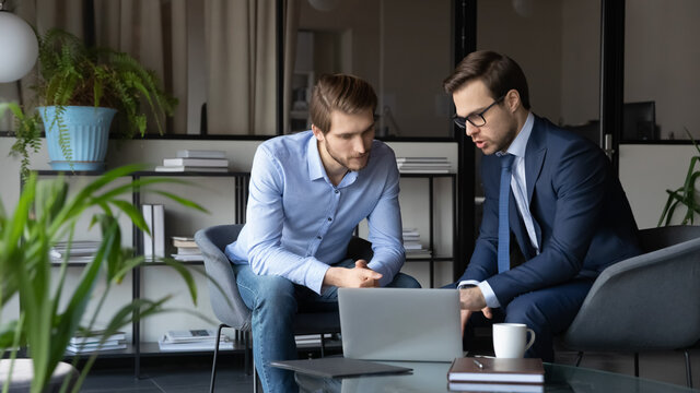Concentrated caucasian businessman in formal suit talk brainstorm cooperate with male colleague at meeting in office, serious men business partners look at laptop discuss financial project together