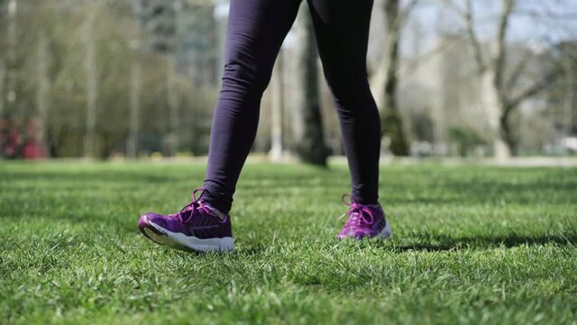 Woman in sportswear walking on grass in park. Female legs and fits wearing tights and sneakers in motion. Cropped shot, closeup. Fitness and outdoor activity concept