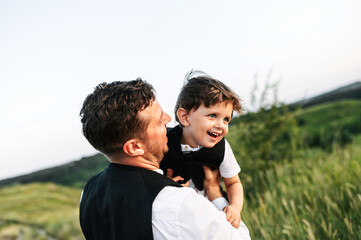 Happy little boy in arms of his dad. Young father and toddler in shirts and suit vests outdoors