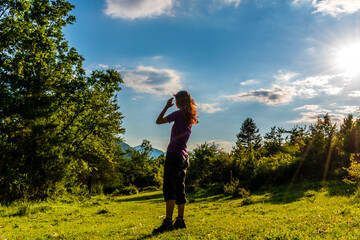 Fototapeta premium A full body shot of an unrecognizable young Caucasian redhead woman standing in a meadow in a forest in the French Alps during sunset