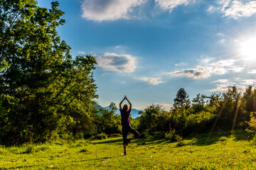 A full body shot of an unrecognizable young Caucasian redhead woman doing yoga in a meadow in a forest in the French Alps during sunset