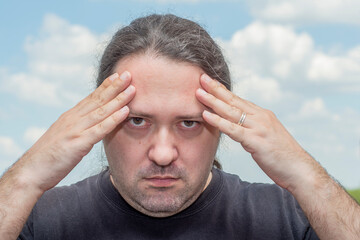 Upset middle-aged adult man with long hair piled in tail pushes his hands to his forehead. Man on a light background.