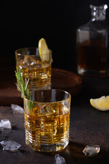 Three glasses of cold whiskey with rosemary, lemon peels on dark brown background. Vertical. Close up.