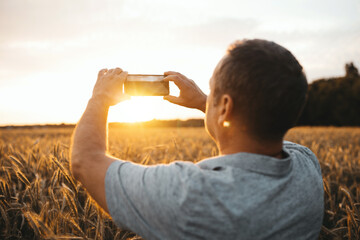Back view of adult farmer taking pictures of sunset or sunrise at golden wheat field. Photographer stand during ripe harvest season. Late summer or early autumn.