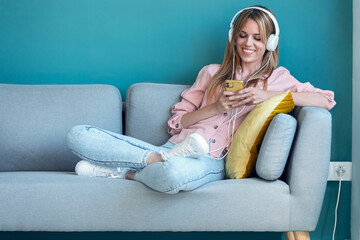 Obraz premium Smiling young woman listening to music with smartphone while sitting on sofa at home.