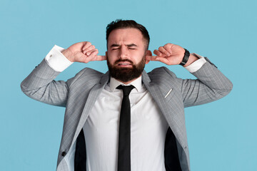 Young businessman stressed by noise, closing both ears over blue background