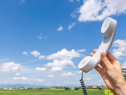 An old white telephone handset is lifted by one hand towards a beautiful blue sky with white clouds with the countryside in the background