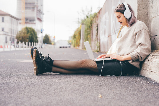 Young woman sitting outdoor using computer listening playlist - Caucasian woman outdoors watching video streaming with laptop and headphones - 5g technology, entertainment, enjoying concept
