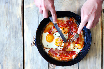 Selective focus. Male hands with a knife and a fork are cutting bacon and eggs in a frying pan. Eggs with bacon. Correct breakfast. The keto diet.