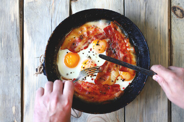 Selective focus. Male hands with a knife and a fork are cutting bacon and eggs in a frying pan. Eggs with bacon. Correct breakfast. The keto diet.