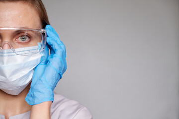 Close up of female doctor or scientist with a medical mask and hands in latex gloves over grey background. She is putting on glasses