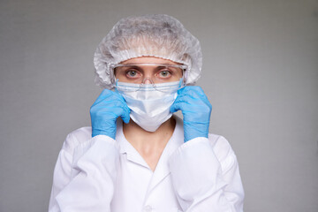 Close up of female doctor or scientist with a medical mask, surgical cap, glasses and hands in gloves over grey background.