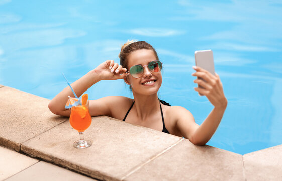 Lovely Caucasian girl taking selfie on smartphone in pool at tropical resort