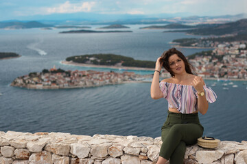 Fototapeta na wymiar Attractive brunette sitting on a stone wall on a warm summer windy day. Fashionable girl posing with the adriatic sea and the small town of primosten in the background