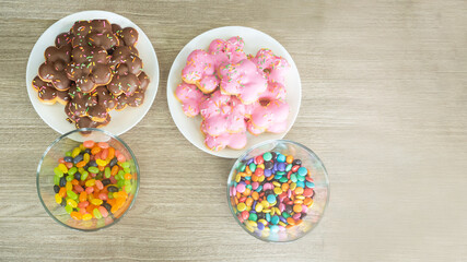 Chocolate and Strawberry glazed donuts in the white plate, colorful jellybeans and round chocolate in the bowls put on wooden table with copy space.