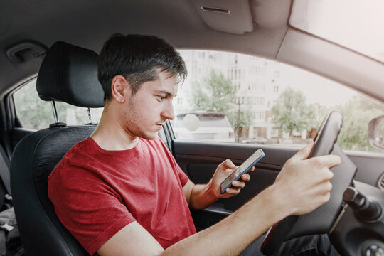 Man Behind The Wheel Of His Car Loses Concentration On The Road Situation And Gets Stuck In A Smartphone Typing SMS In A Chat. Risk Of A Dangerous Road Accident