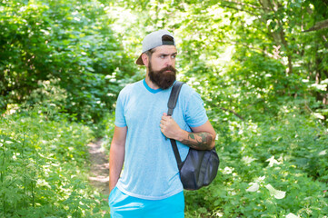Naklejka na ściany i meble bearded man look confidently. brunette bearded man wear cap in forest on background of trees. brutal brunette bearded man with backpack. mature guy with beard in wood. nice summer day