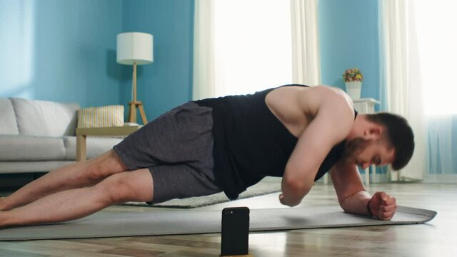 Strong athletic man is doing exercises standing in side elbow plank at home, using stopwatch on a gadget, making efforts to maintain his perfect body shape, Zoom in, Slow motion.