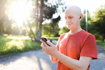 Smiling old man in casual wear and white headphones listening music from his smartphone in the park.