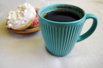 a large turquoise mug of hot coffee against a brown plate with round doughnuts with pink icing and a white one with marshmallows on a beige tablecloth
