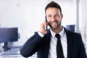 Portrait shot of busy businessman using his mobile phone and talking with somebody while standing in the office.