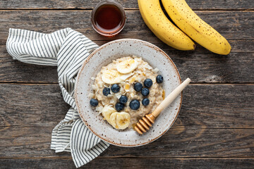 Oatmeal porridge with honey, blueberries and banana in bowl on rustic wooden table, top view