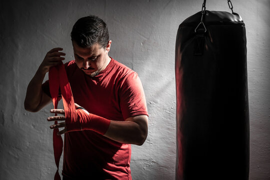 Boxeador joven principiante con camiseta roja poniendose cinta en las manos junto a un saco negro en su garaje con guantes de boxeo negros, iluminado con luz intensa de flash y fondo negro