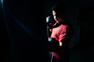 Fototapeta na wymiar Boxeador joven principiante con camiseta roja golpeando un saco negro en su garaje con guantes de boxeo negros, iluminado con luz intensa de flash y fondo negro