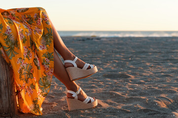 Legs of a female in a yellow dress with floral patterns and elegant white wedges at the beach