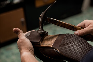 Male cobbler holding shoe and nailing a heel with hummer. Close-up on hands of professional shoemaker. - obrazy, fototapety, plakaty