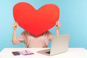 Woman hiding face behind huge red paper heart, symbol of love, romantic feelings, using anonymous dating service on laptop, online chat and website. indoor studio shot isolated on blue background